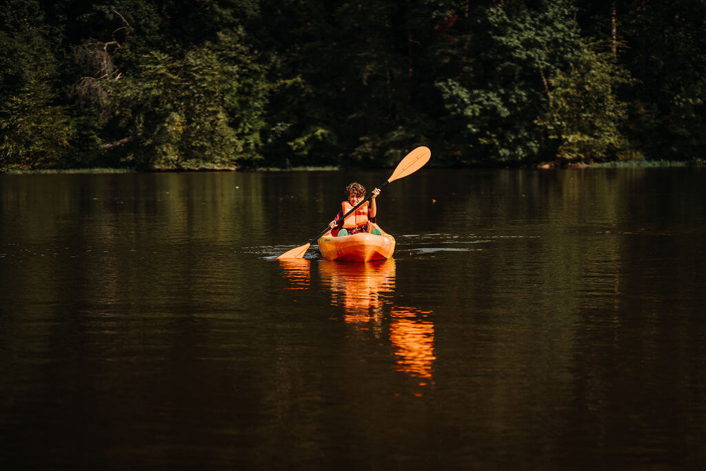 A person wearing an orange life jacket paddles a yellow kayak on a calm lake surrounded by dense green trees. The water reflects the kayak and the greenery, creating a serene and peaceful scene.