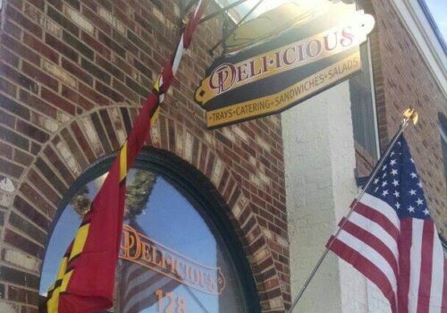 A brick building with an arched window and two flags hanging above the entrance. One flag is the American flag, and the other is a state or regional flag. A sign above reads 