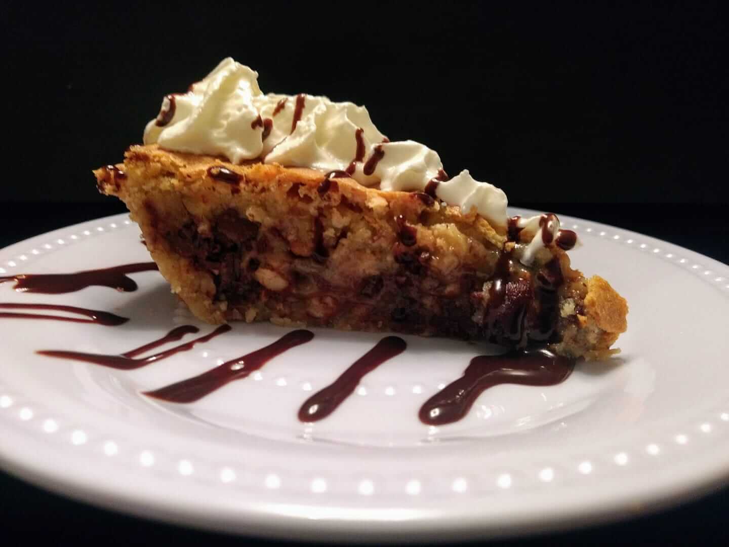 A slice of pie with a golden crust, filled with what appears to be pecans and chocolate chips, topped with whipped cream. The pie is served on a white plate drizzled with chocolate sauce against a black background.