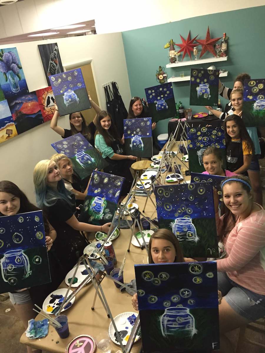 A group of people, mostly young women, are seated around a table at a painting class. Each person holds up their completed canvas of a robot in a blue and green background. The room has a casual atmosphere with art supplies and colorful decor visible.