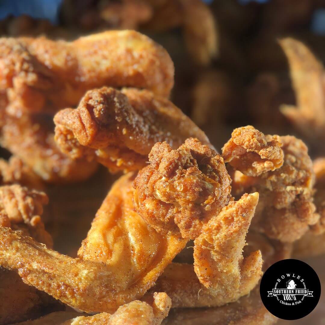 Close-up of crispy, golden-brown fried chicken wings, showcasing their crunchy texture. A black and white logo in the bottom right corner displays 