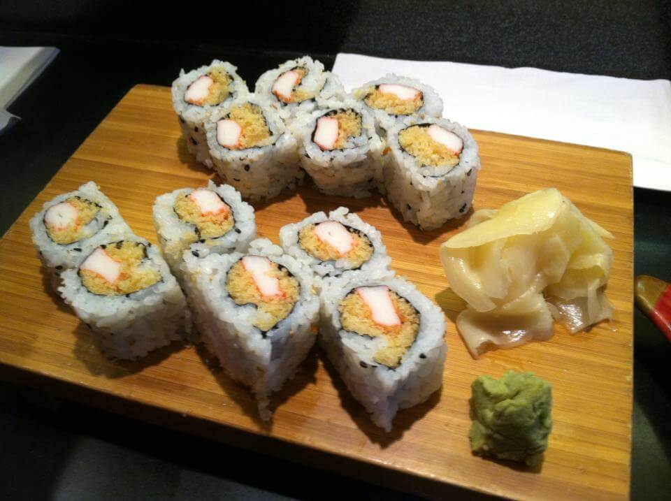 A wooden board with eight sushi rolls, each filled with crab meat and a crunchy tempura center, topped with sesame seeds. Next to the rolls are servings of pickled ginger and a small mound of wasabi.