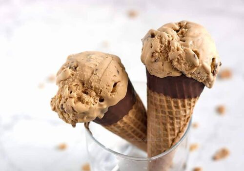 Two chocolate-dipped waffle cones filled with scoops of creamy, caramel-colored ice cream studded with pieces of toffee, standing upright in a clear glass on a white background. Crumbs are scattered around. Image