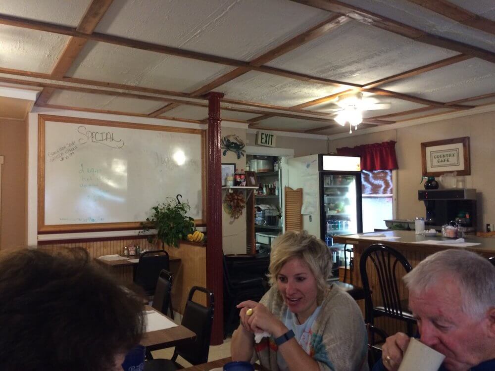 Inside a cozy café with wooden ceiling beams and whiteboard menu, a woman with short blonde hair and a man with gray hair sit at a table, engaged in conversation. The man drinks from a mug, while another person's head is partially visible in the foreground.