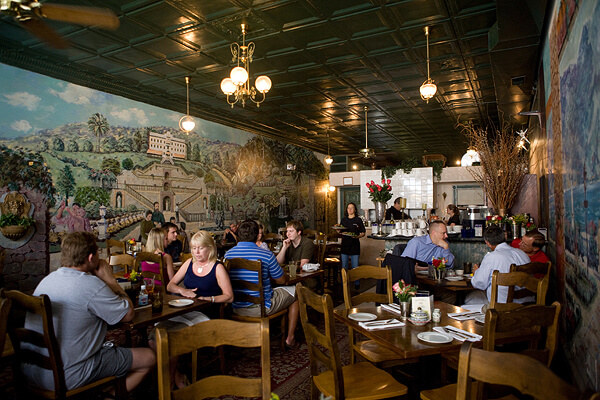 A cozy restaurant with wooden furniture, colorful murals on the walls depicting scenic landscapes, and decorative lighting. Patrons are seated at tables, enjoying their meals. A few waitstaff are seen serving food and drinks, contributing to a lively atmosphere.