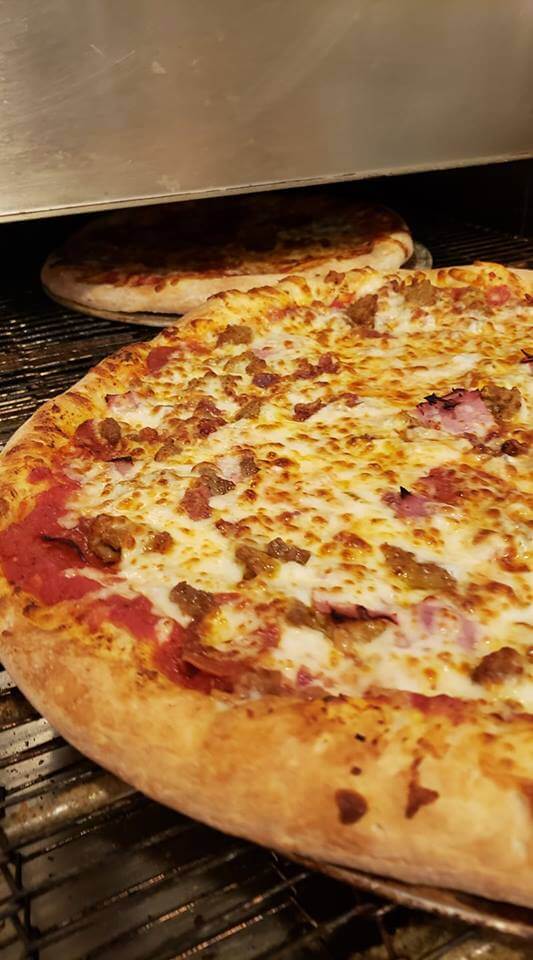A close-up view of two large pizzas baking in an oven. The foreground pizza has a golden-brown crust and is topped with melted cheese, bits of meat, and other toppings. Another pizza is visible baking on the rack above.