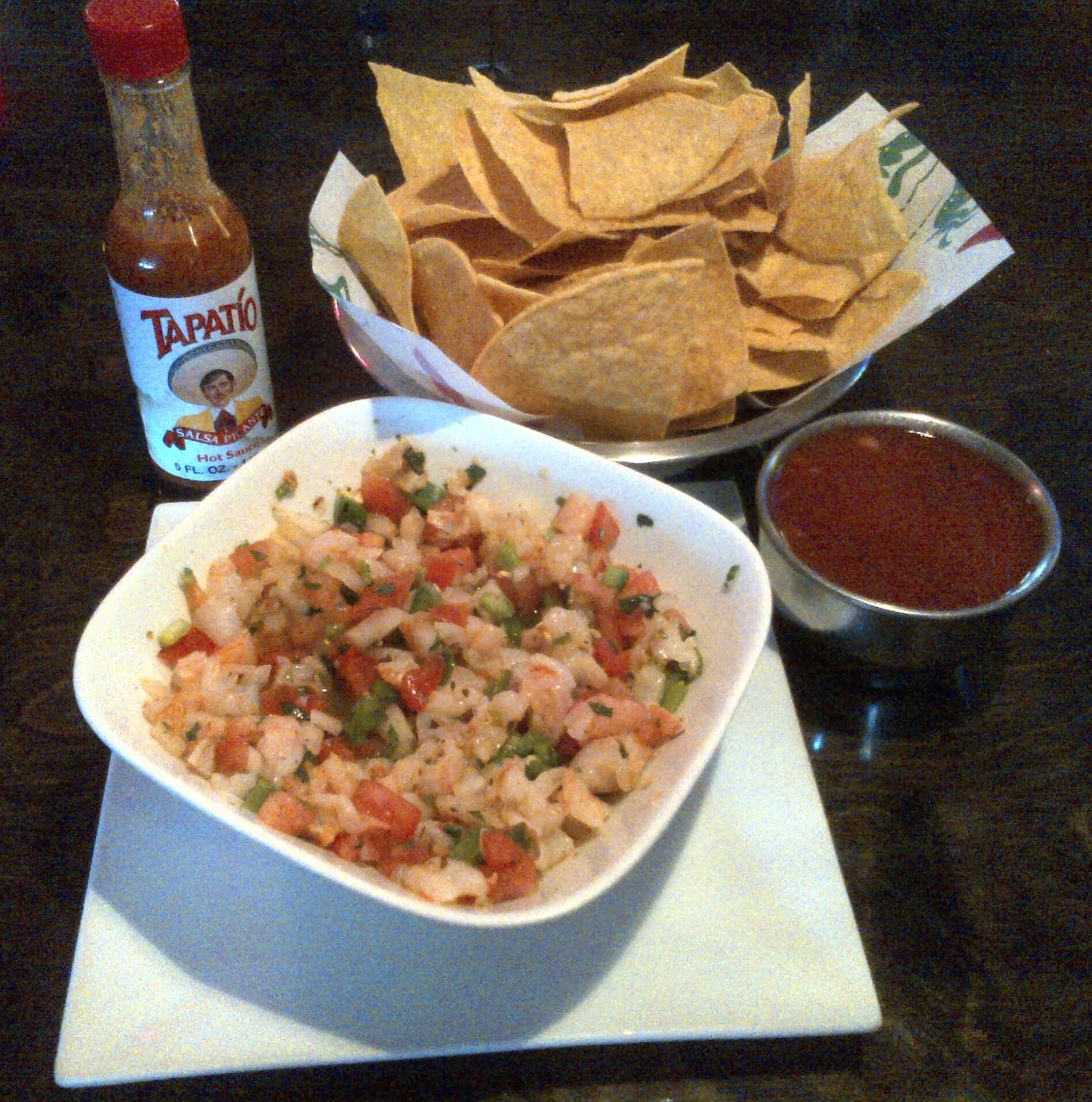 A bowl of tortilla chips next to a metal cup filled with salsa, a square white bowl of pico de gallo, and a bottle of Tapatío hot sauce, all placed on a dark wooden table.