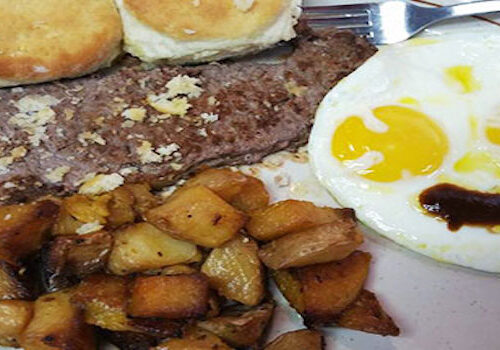 A plate featuring sunny side up eggs with a smiley face made of sauce, a beef patty, roasted potato cubes, and two biscuits. A fork is placed next to the eggs. Image