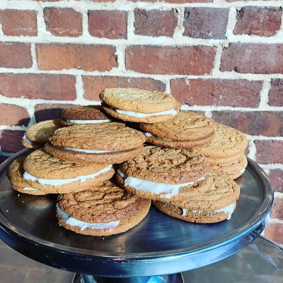 A stack of homemade oatmeal cream pies sits on a round, metallic tray. The cookies have a rough texture, and each one is generously filled with a layer of white cream. The tray is placed in front of a rustic brick wall.