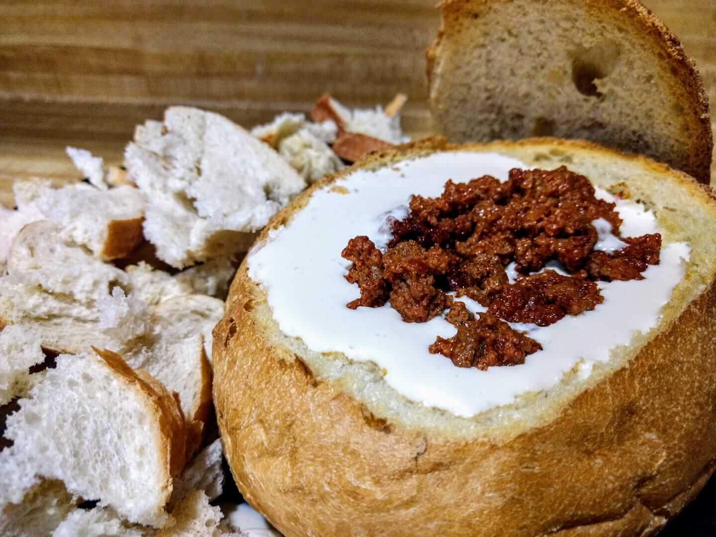 A bread bowl filled with creamy white dip topped with crumbled chorizo. The bread bowl sits on a wooden surface, surrounded by torn pieces of bread. The bread has a crispy crust and a soft, fluffy interior.