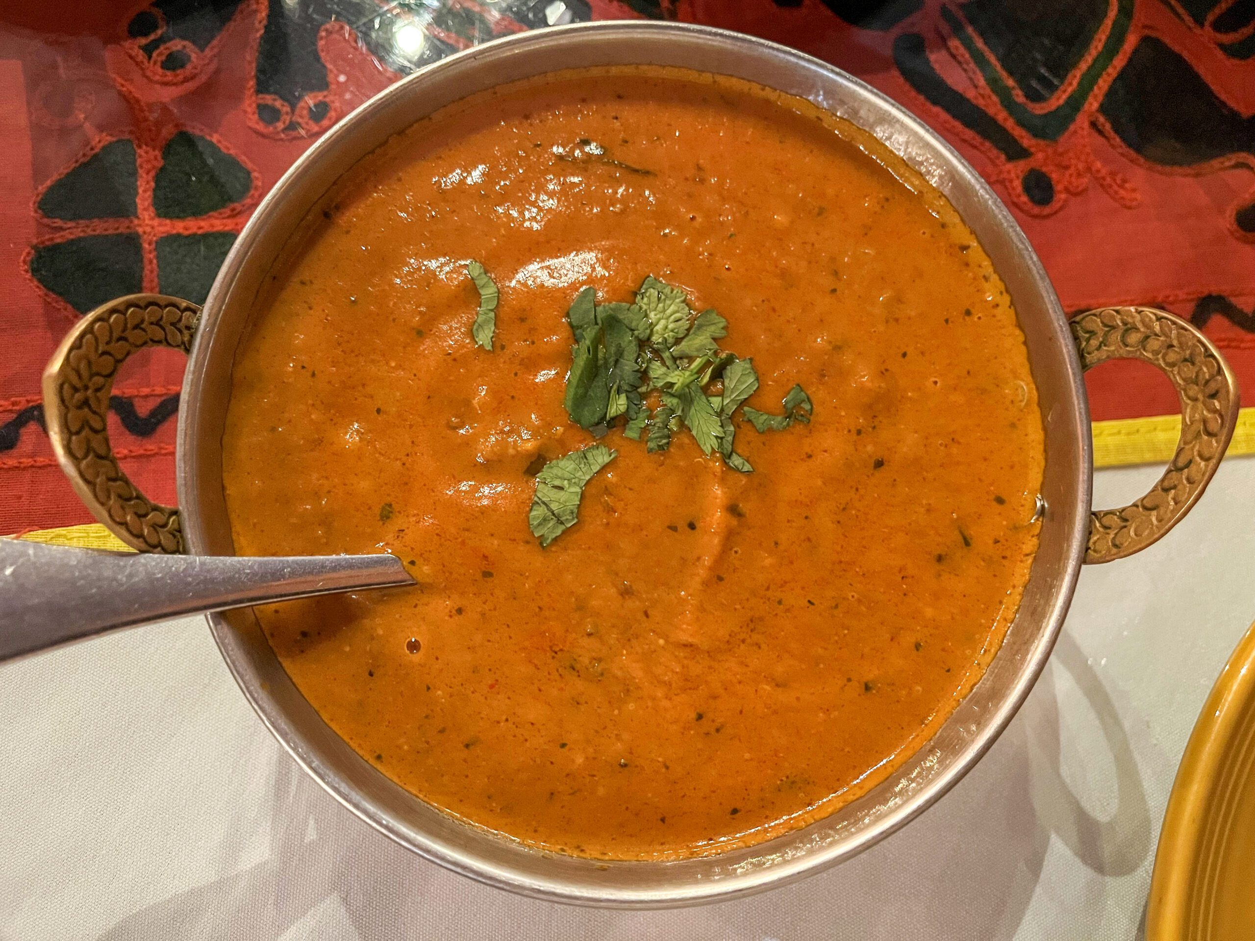 A bowl of creamy orange-red tomato soup garnished with fresh cilantro, served in a decorative bowl with two handles. A spoon rests inside the bowl. The background features a red and black patterned tablecloth.