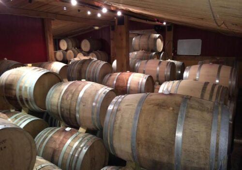 A room with wooden barrels stacked on their sides, arranged in rows. The barrels are secured with metal bands and are placed on wooden supports. The room has wooden walls and ceilings with small lights hanging from the ceiling. Image