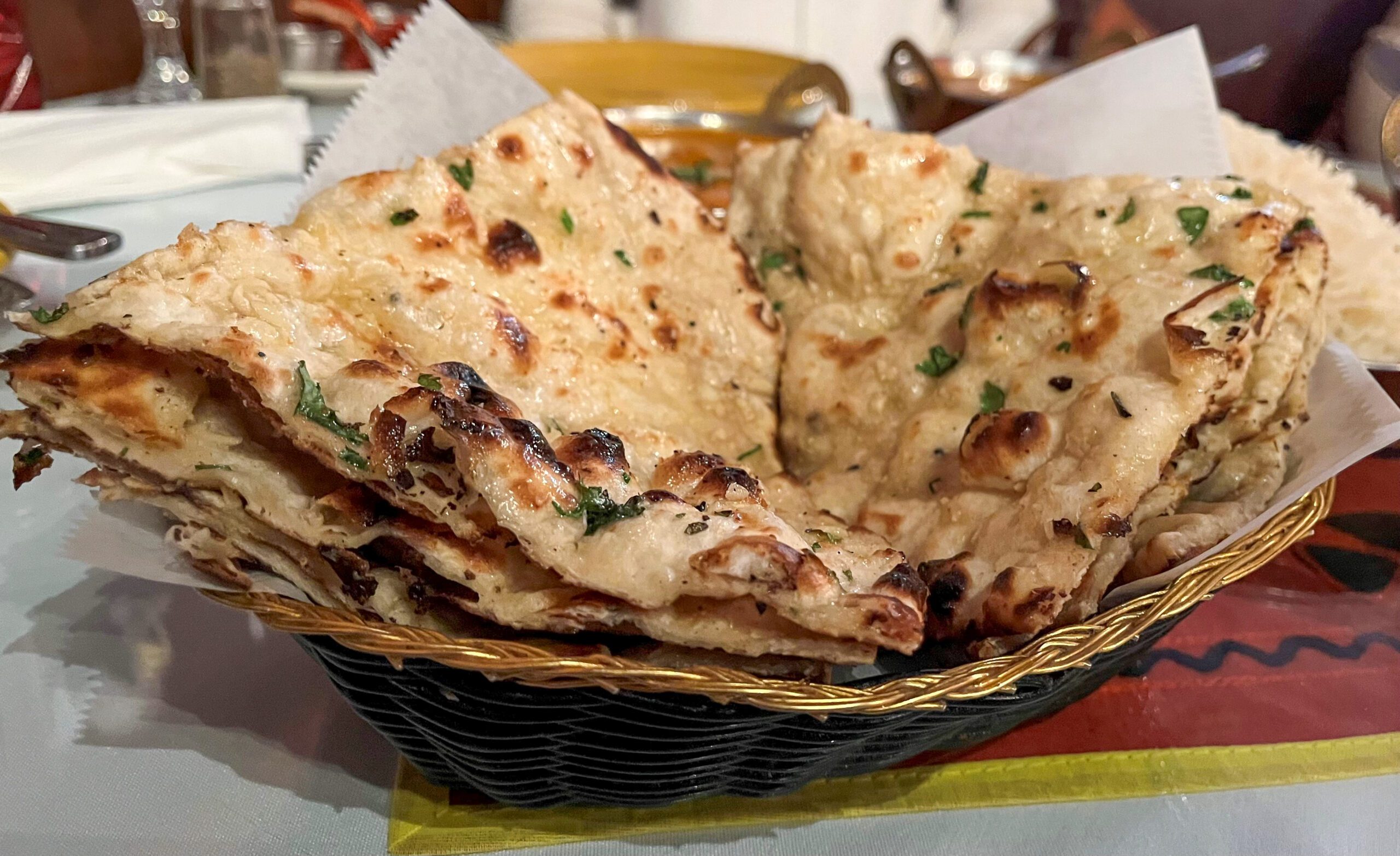 A basket filled with sliced pieces of naan bread garnished with fresh herbs. The naan is lightly toasted with some charred, crispy edges, indicating it has been cooked in a tandoor. The background shows a blurred dining setting with plates and utensils.