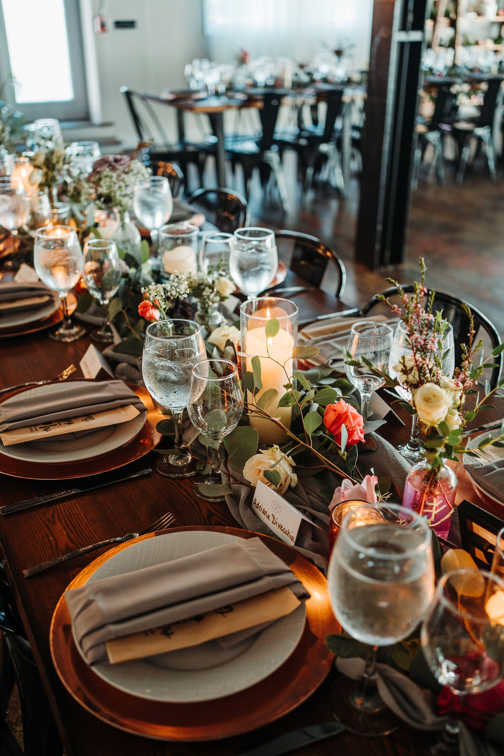 A decorated dining table set up for an event. The table features neatly arranged plates and silverware, water glasses, candles, and floral arrangements. The background shows additional tables set similarly with a warm, inviting atmosphere.