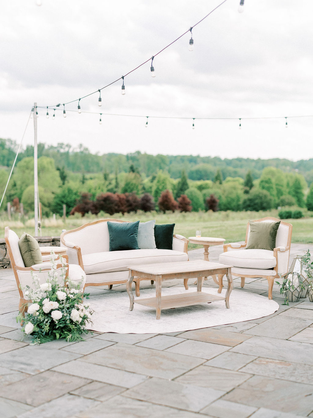 An outdoor seating area features a white cushioned sofa with black pillows, two matching armchairs with green pillows, and a wooden coffee table on a round white rug. Decorative string lights hang above, with a lush green landscape in the background.