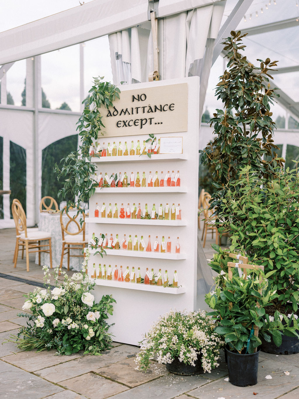A decorative white board at an outdoor event, adorned with greenery and colorful drink bottles. The board reads 
