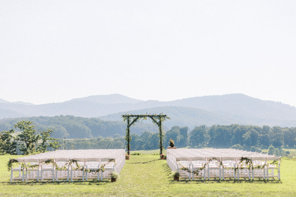 An outdoor wedding ceremony setup in a grassy field with mountains in the background. White folding chairs are arranged in rows facing a wooden arbor adorned with greenery. The sky is clear, and trees line the horizon.