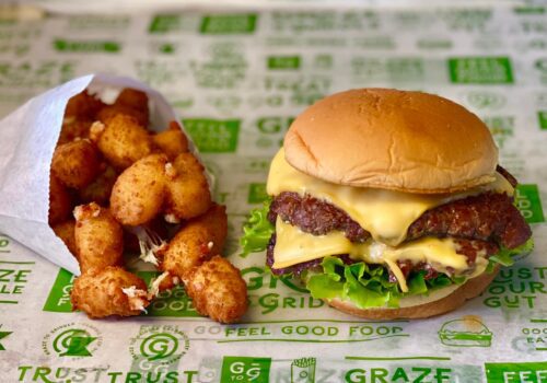 A cheeseburger with lettuce and double patties sits on a wrapper with green text. Next to it is a side of fried cheese curds in a white paper bag. The background wrapper features various green phrases and logos related to the food brand. Image