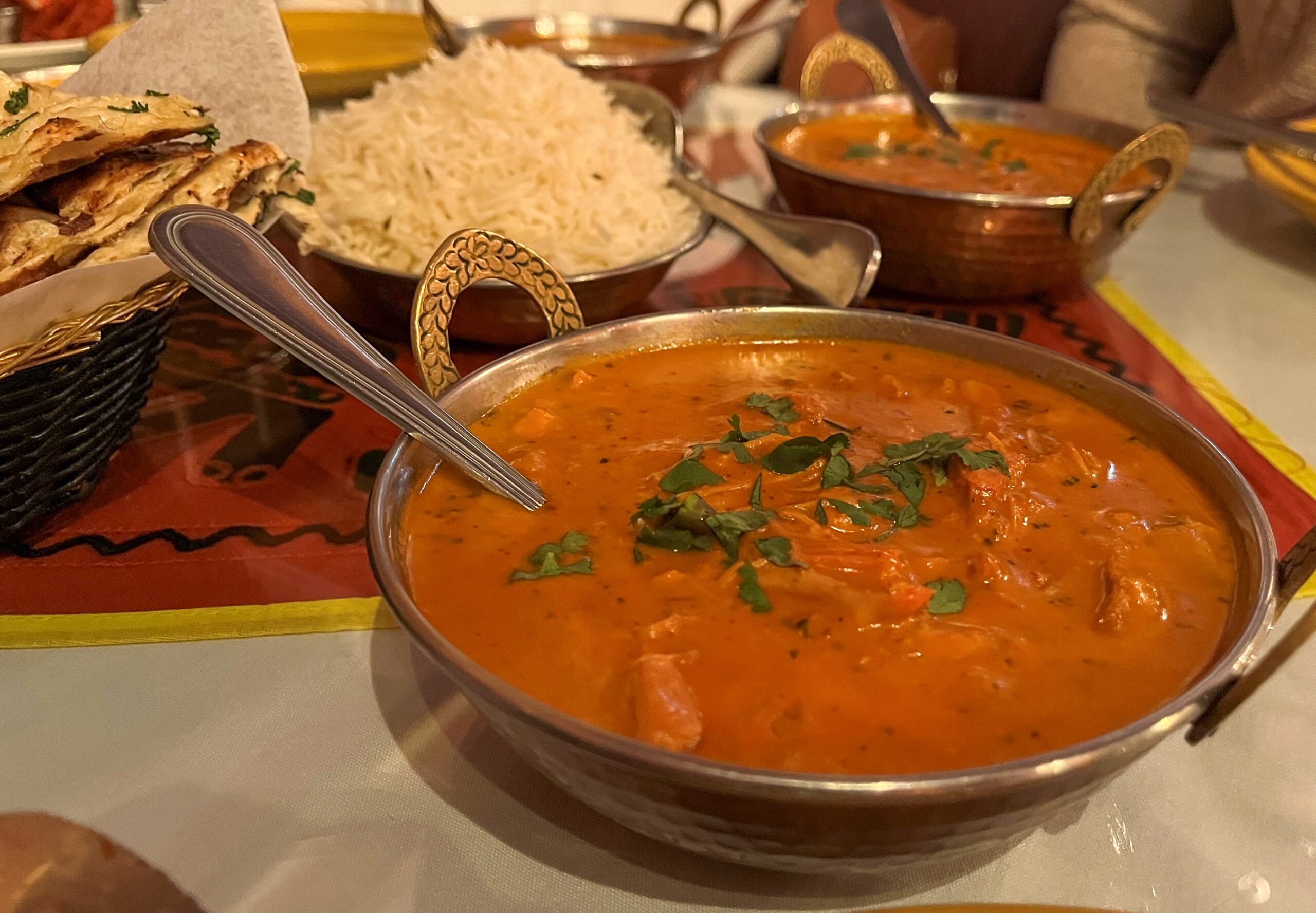A close-up of a meal featuring a bowl of curry garnished with cilantro, a bowl of white rice, and naan bread in a basket. The dishes are placed on a table with a red and yellow patterned tablecloth. Spoons are in the curry and rice.