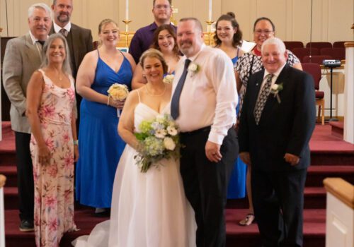 A bride and groom stand in the center wearing wedding attire, surrounded by a group of smiling people in formal clothing. They are inside a church with a stained glass window and lit candles in the background. Image