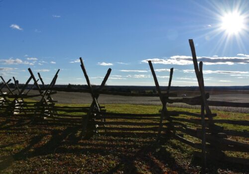 A landscape view featuring a rustic wooden fence with angled posts under a bright blue sky. The sun is shining brightly from the top right corner, casting long shadows on the grassy field below. Sparse clouds are scattered across the horizon. Image
