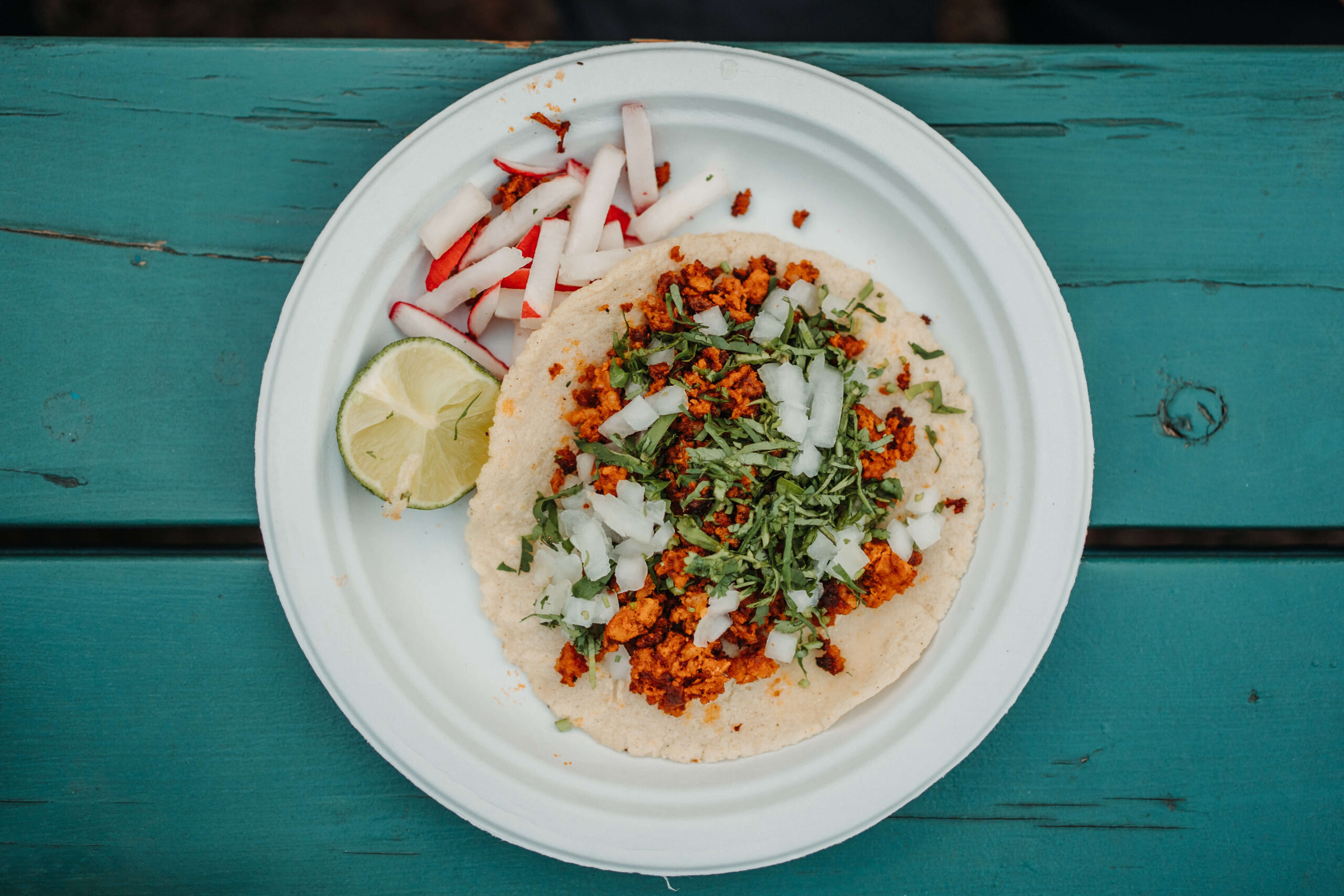 A taco filled with seasoned ground meat, chopped onions, and cilantro on a white plate. Garnished with a lime wedge and sliced radishes, the plate rests on a teal wooden surface.