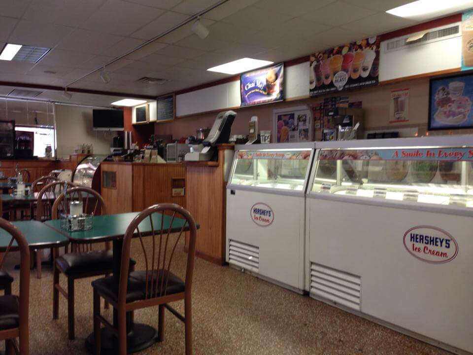 A brightly lit ice cream parlor featuring a Hershey's Ice Cream display freezer. The counter in the background has an assortment of toppings and an ice cream maker. Several empty tables with green tops and wooden chairs are in the foreground.
