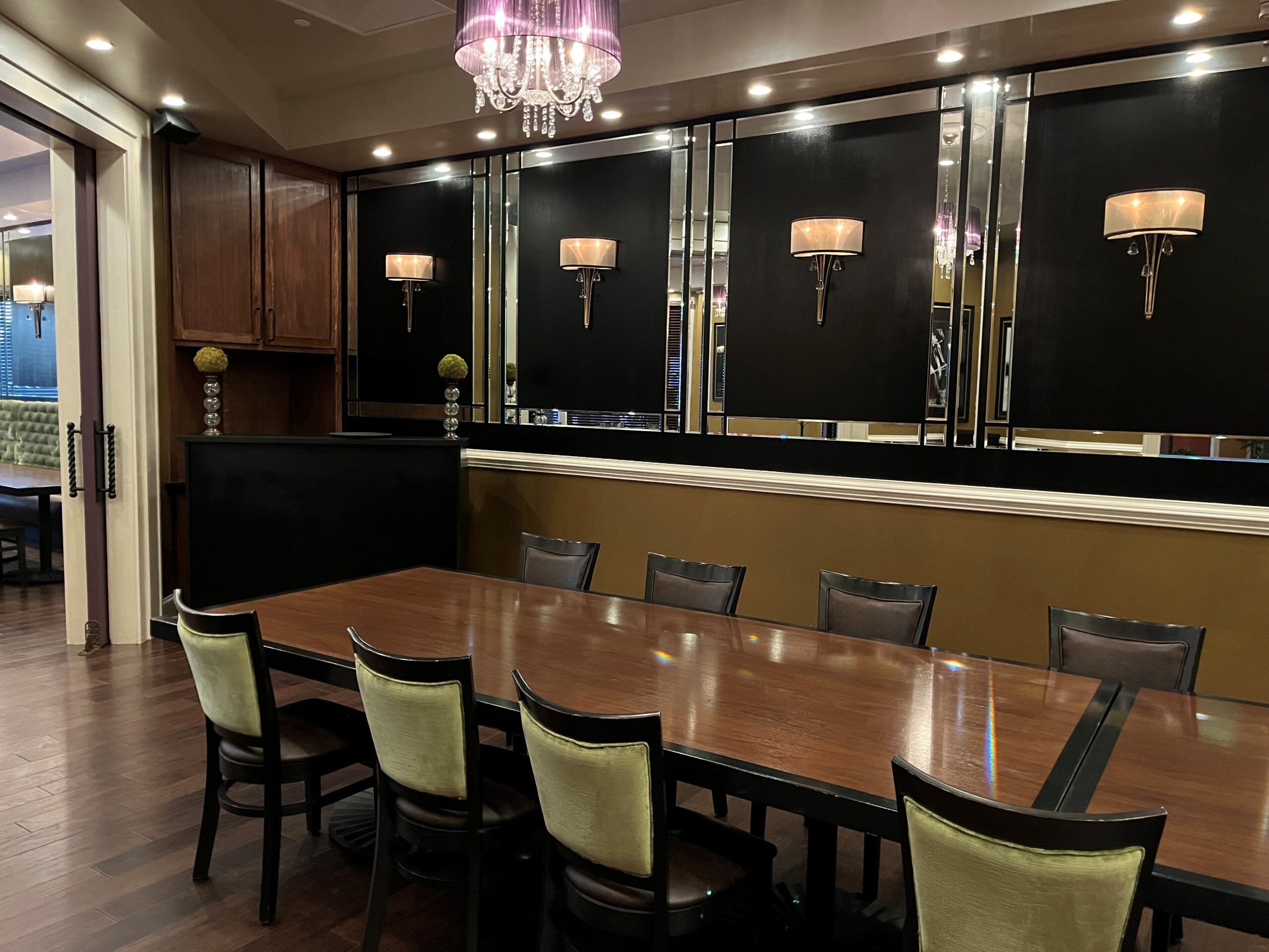 A modern dining room featuring a long wooden table surrounded by eight chairs. The room is adorned with wall-mounted light fixtures, decorative mirrors, and a hanging chandelier. The decor includes black, gold, and wooden accents, creating an elegant ambiance.