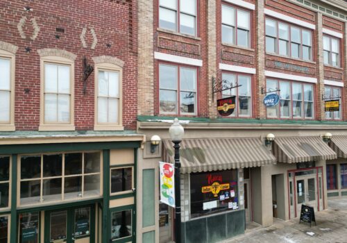 A streetscape featuring two-story brick buildings with multiple awnings and signs for various businesses, including a martial arts studio. The buildings have large windows and decorative brickwork. A lamppost with colorful banners stands on the sidewalk. Image
