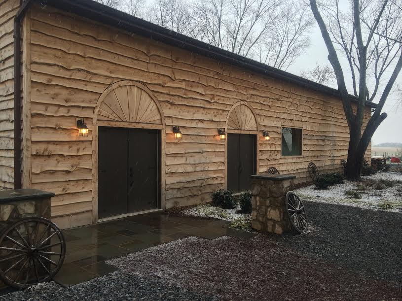 A rustic, wooden building with two double doors, flanked by four warm-lit wall lamps. Snow is lightly covering the ground and the nearby trees, with some stone and wagon wheel decorations enhancing the building's quaint, rural appearance.