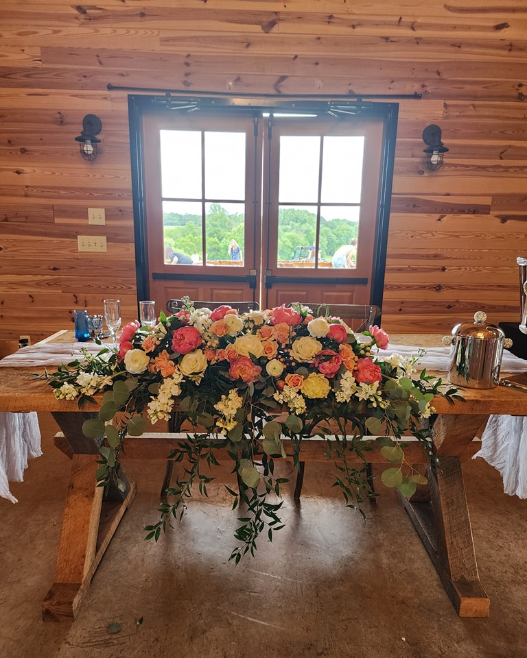 A rustic wooden table decorated with a vibrant bouquet of colorful flowers sits in front of a large window with a view of greenery outside. The table has glasses, a champagne bucket, and is set against a wooden-paneled wall with small light fixtures.