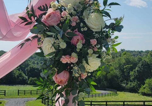 A rustic wooden wedding arch is adorned with pink and white roses, baby's breath, green foliage, and draped with pink tulle in a scenic outdoor setting with a lush green field and split-rail fence under a clear blue sky. Image