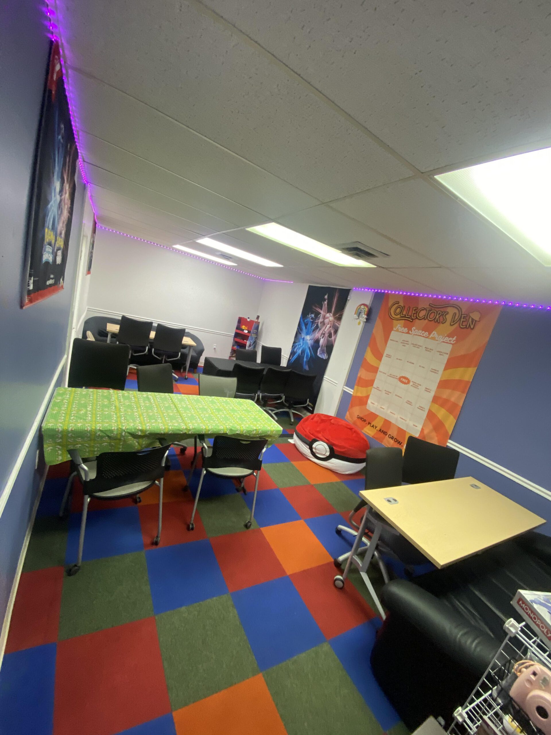 A room with colorful checkered flooring, blue walls, and white ceiling. It has tables with chairs, a sofa, posters on the walls, and string lights along the ceiling. There is a bright chart on the right wall and a red bean bag on the floor.
