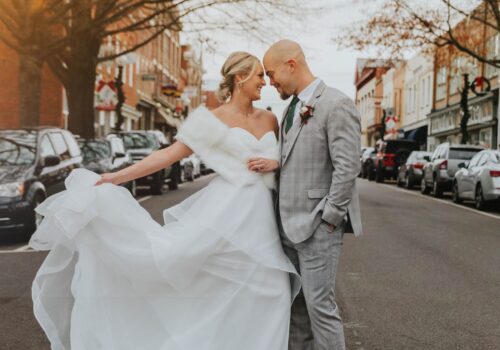 A bride in a white gown and a groom in a gray suit stand closely together on a street lined with parked cars and buildings. The bride holds up part of her dress, and both are smiling warmly at each other. Leafless trees line the street, adding a wintery atmosphere. Image
