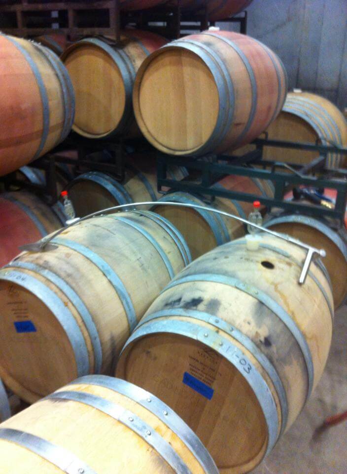 A stack of wooden wine barrels organized on metal racks, some with airlocks inserted. The barrels have metal bands and small labels, and the surrounding area appears to be part of a winery or cellar. The image is slightly blurry.