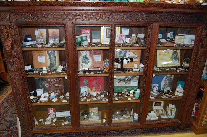 A wooden cabinet with intricately carved details and glass doors houses various collectibles. The shelves display books, small figurines, jewelry, and other memorabilia. The cabinet stands on a patterned rug in a room with various other antiques.