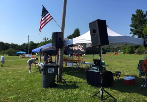 Outdoor event setup with a U.S. flag on a pole, a DJ booth under a tent displaying 