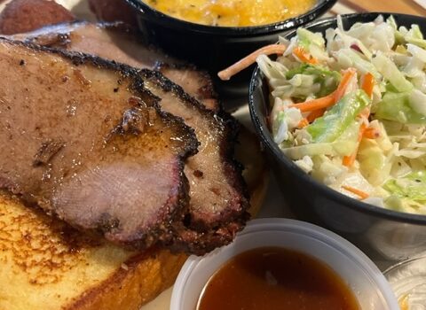 A plate of barbecue food featuring two slices of smoked brisket on a piece of toast, a side of coleslaw, a small bowl of cheesy potatoes, and a container of barbecue sauce. The meal is presented on a ceramic plate with utensils visible in the background. Image