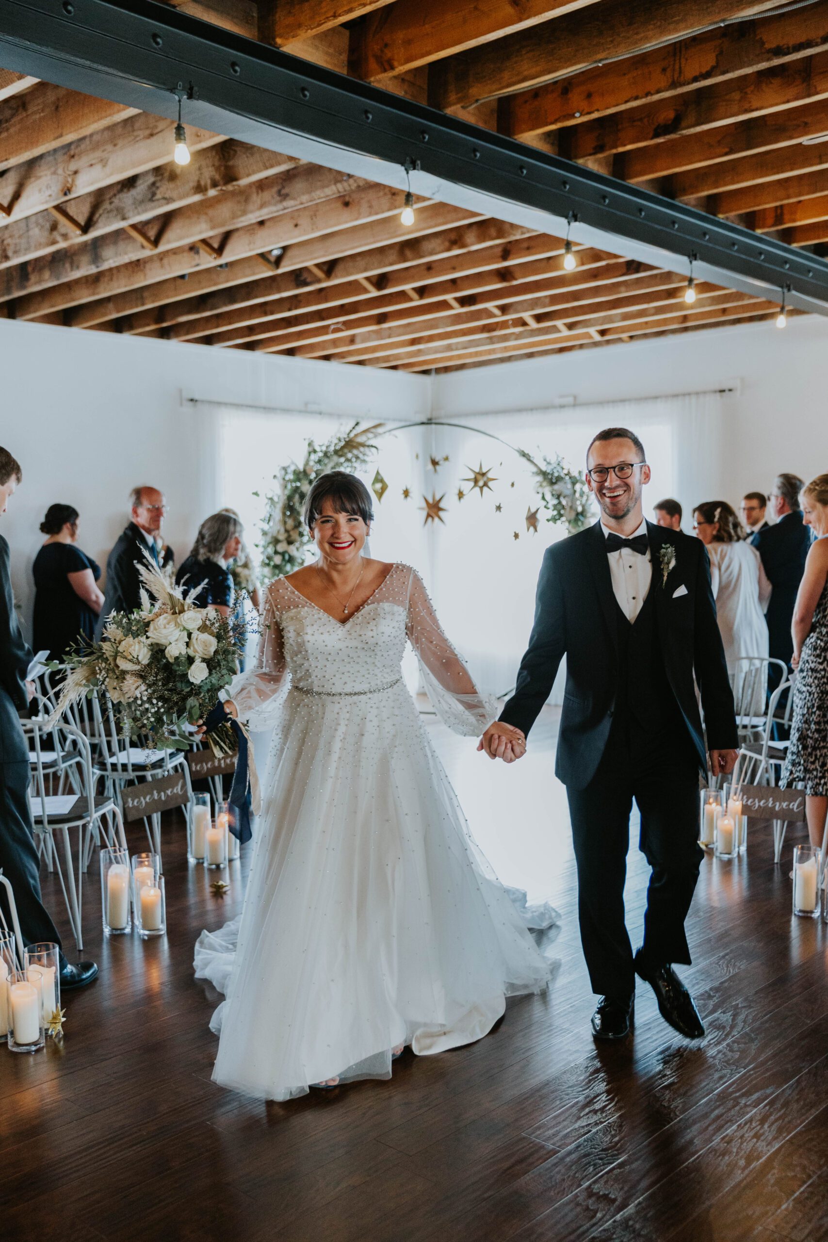 A bride and groom walk down the aisle of a stunning wedding and event venue in Culpeper, Virginia. They smile, holding hands—the bride in a white gown with long sleeves, the groom in a black tuxedo. Guests are seated on either side, while flowers and candles adorn the aisle beneath a rustic wooden ceiling.