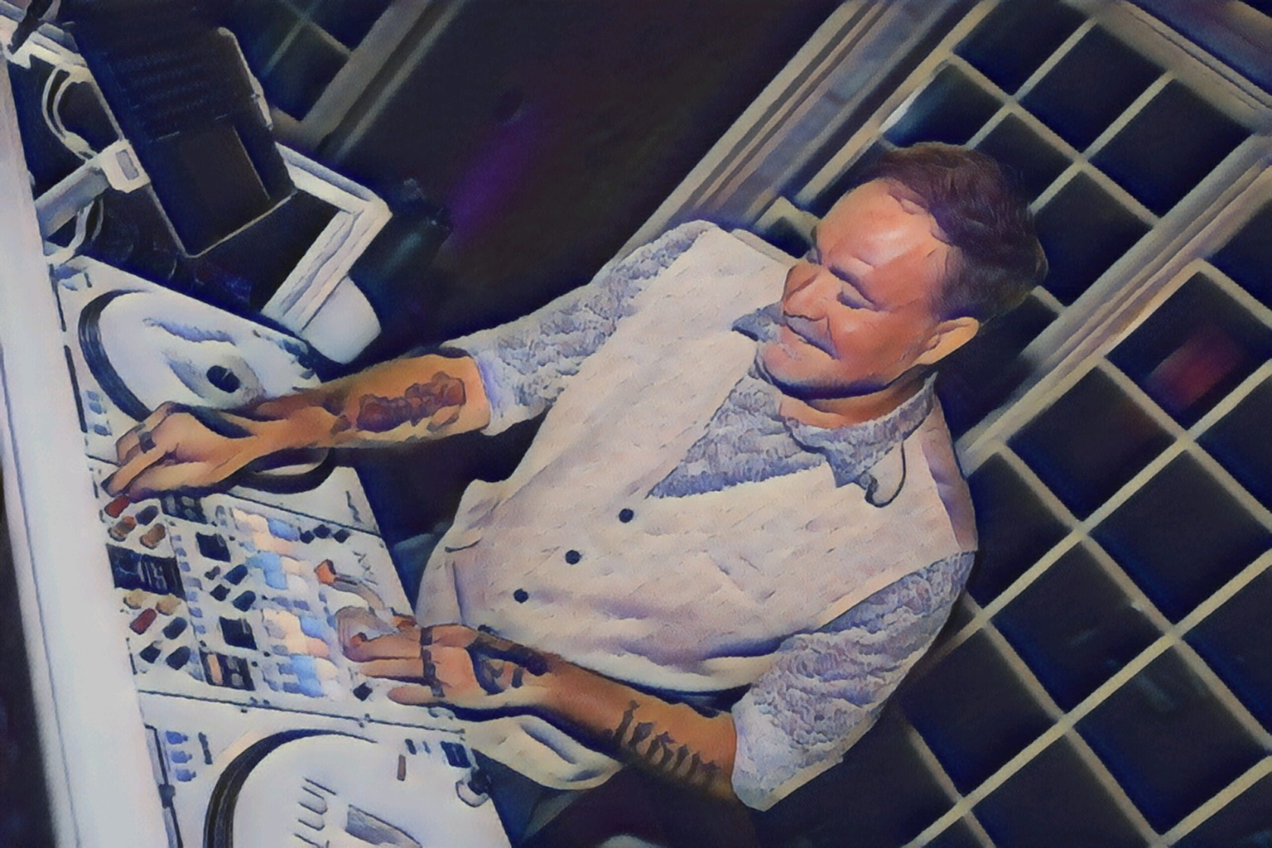 A person with short hair and tattoos stands at a DJ booth, mixing music on a digital turntable. They are wearing a light-colored vest over a patterned shirt and are smiling as they work. The background features a large window with multiple panes.