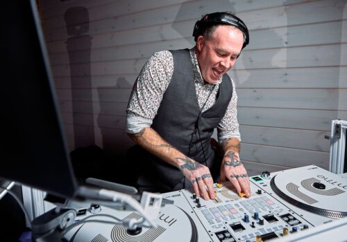 A DJ is energetically mixing music on a set of turntables. He is wearing a grey vest over a patterned shirt, headphones, and has multiple tattoos on his arms and hands. The background features light-colored wooden panels. Image