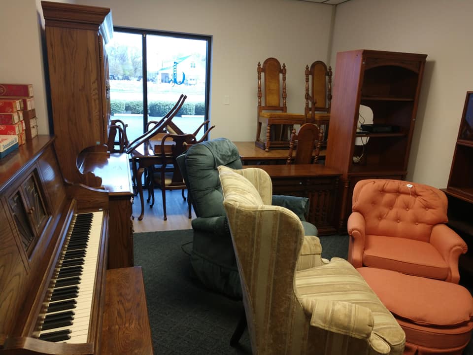 A room filled with various furniture items including a wooden piano, armchairs, cabinets, and tables. Several pieces are stacked or arranged closely together. A window in the background shows an outdoor scene with trees and a blue sky.
