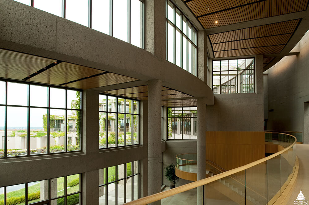 Interior of a modern building with large windows, concrete pillars, and wooden ceilings. The space is open and well-lit, with a winding staircase and greenery visible outside. Light streams in, creating a bright and airy atmosphere.