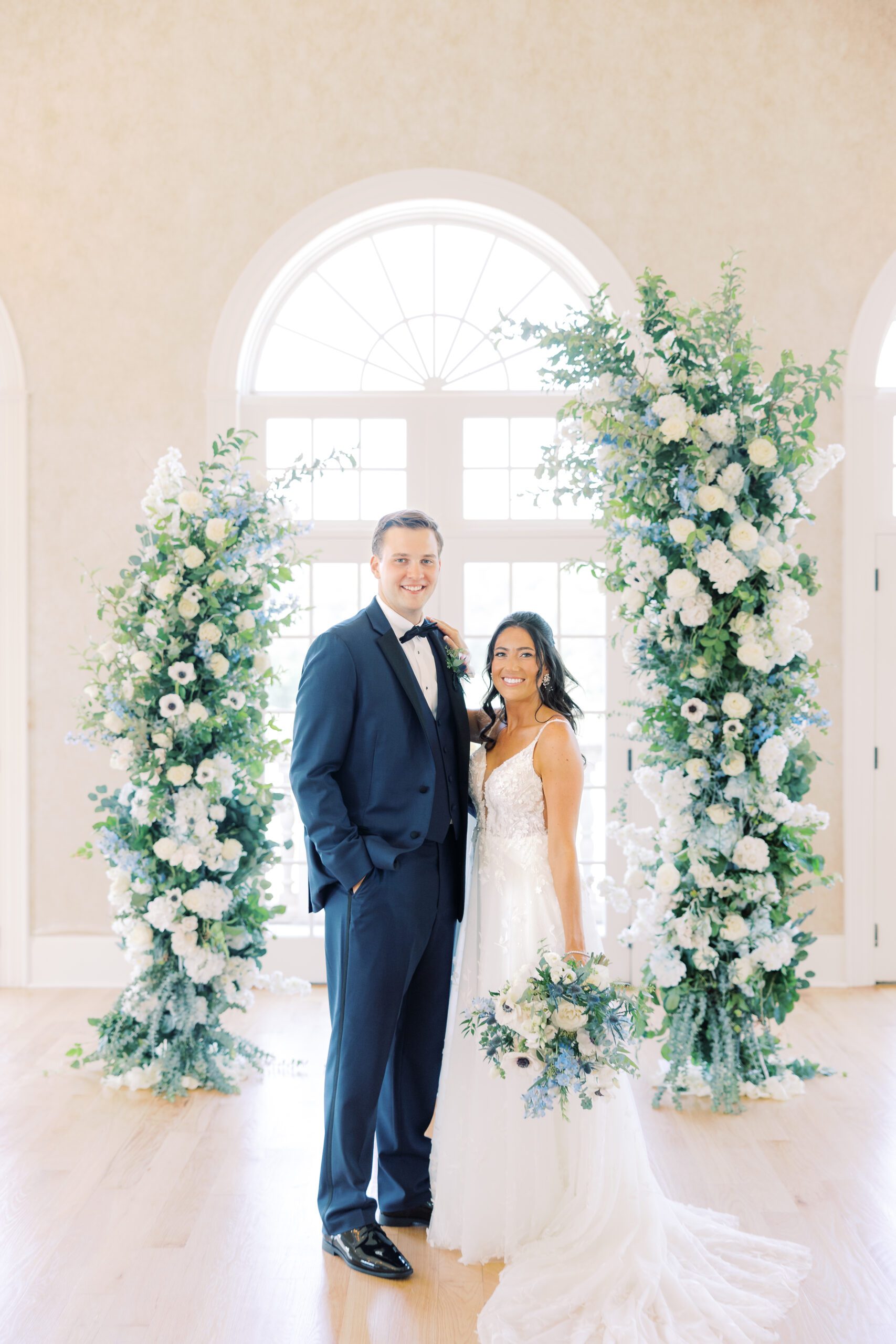 A bride in a white gown and groom in a navy blue suit smile while standing before a large arched window adorned with floral arrangements. The elegant setting features soft lighting, green and white flowers, and a light wooden floor, creating a bright and airy atmosphere.