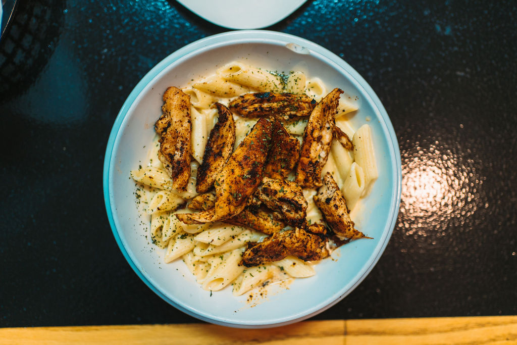 A bowl of penne pasta topped with grilled chicken strips, seasoned with herbs and spices, served on a glossy black surface. The pasta appears to have a creamy white sauce.