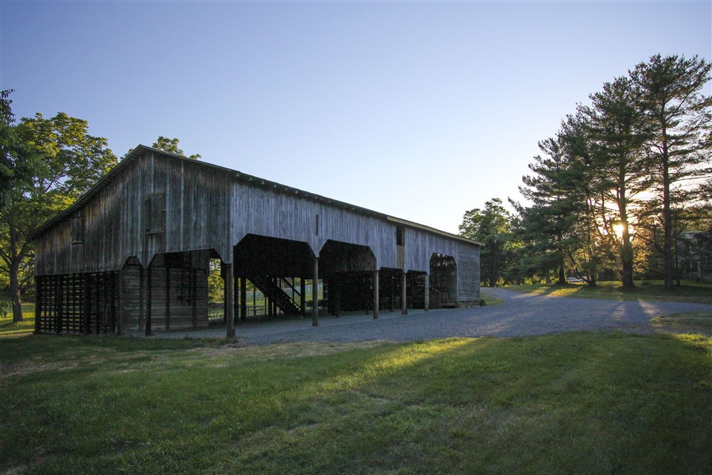 A large, rustic wooden barn stands in a grassy field surrounded by trees during sunrise or sunset. The light casts long shadows on the ground, illuminating the structure and parts of the nearby landscape with a warm glow. A gravel path curves around the barn.