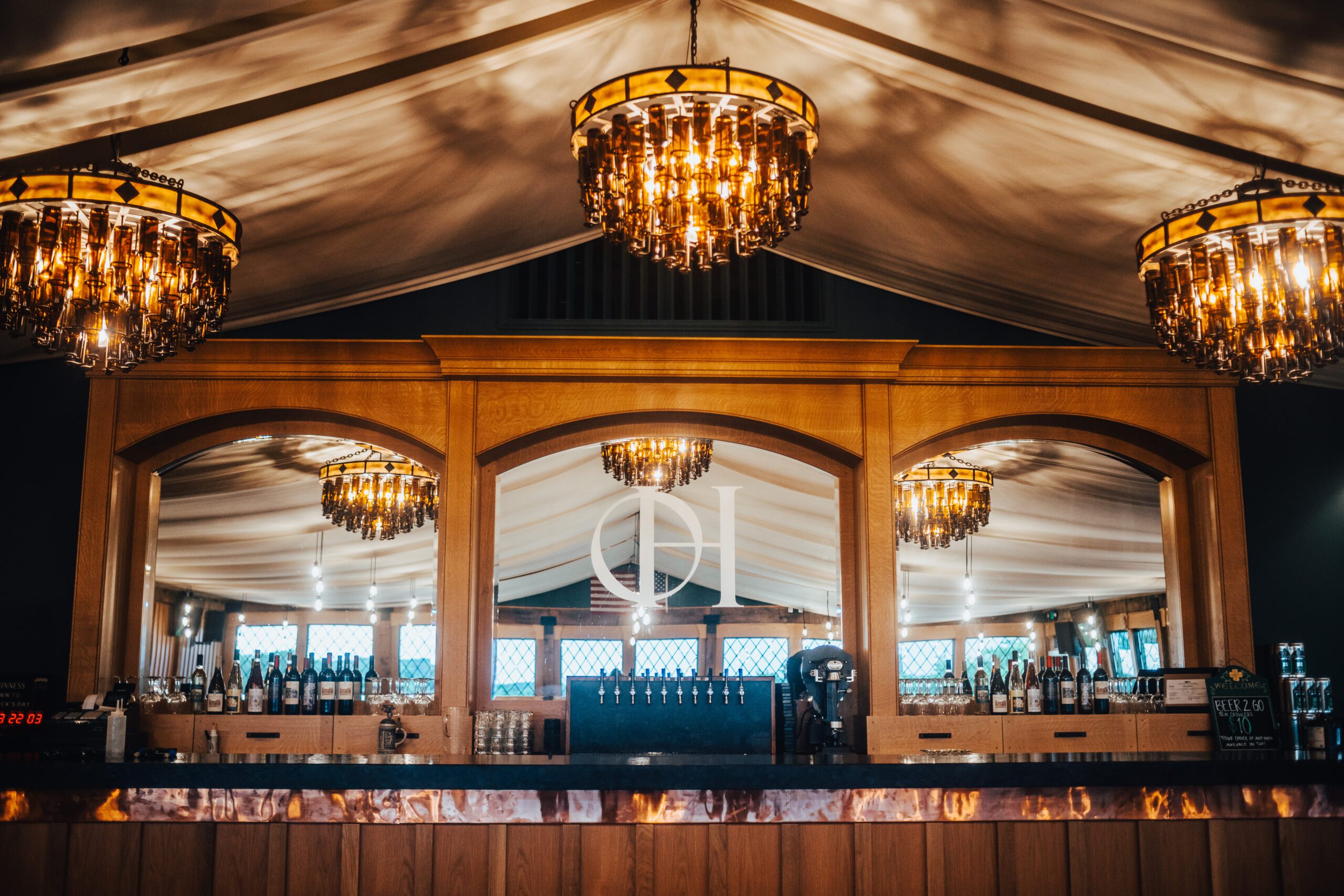 A luxurious bar with three ornate chandeliers reflecting in large mirrors behind a wooden bar counter. The ceiling is draped with white fabric, and the bar is stocked with various bottles and glassware. The ambiance is elegant and warm.