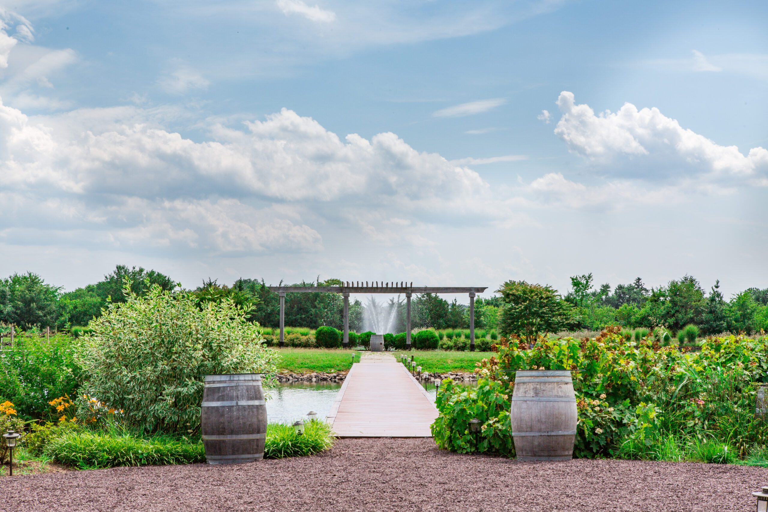 A scenic garden pathway flanked by wine barrels and lush greenery leads to a wooden pergola in the distance. The sky is partly cloudy, and a fountain is visible beyond the pergola, surrounded by vibrant vegetation.
