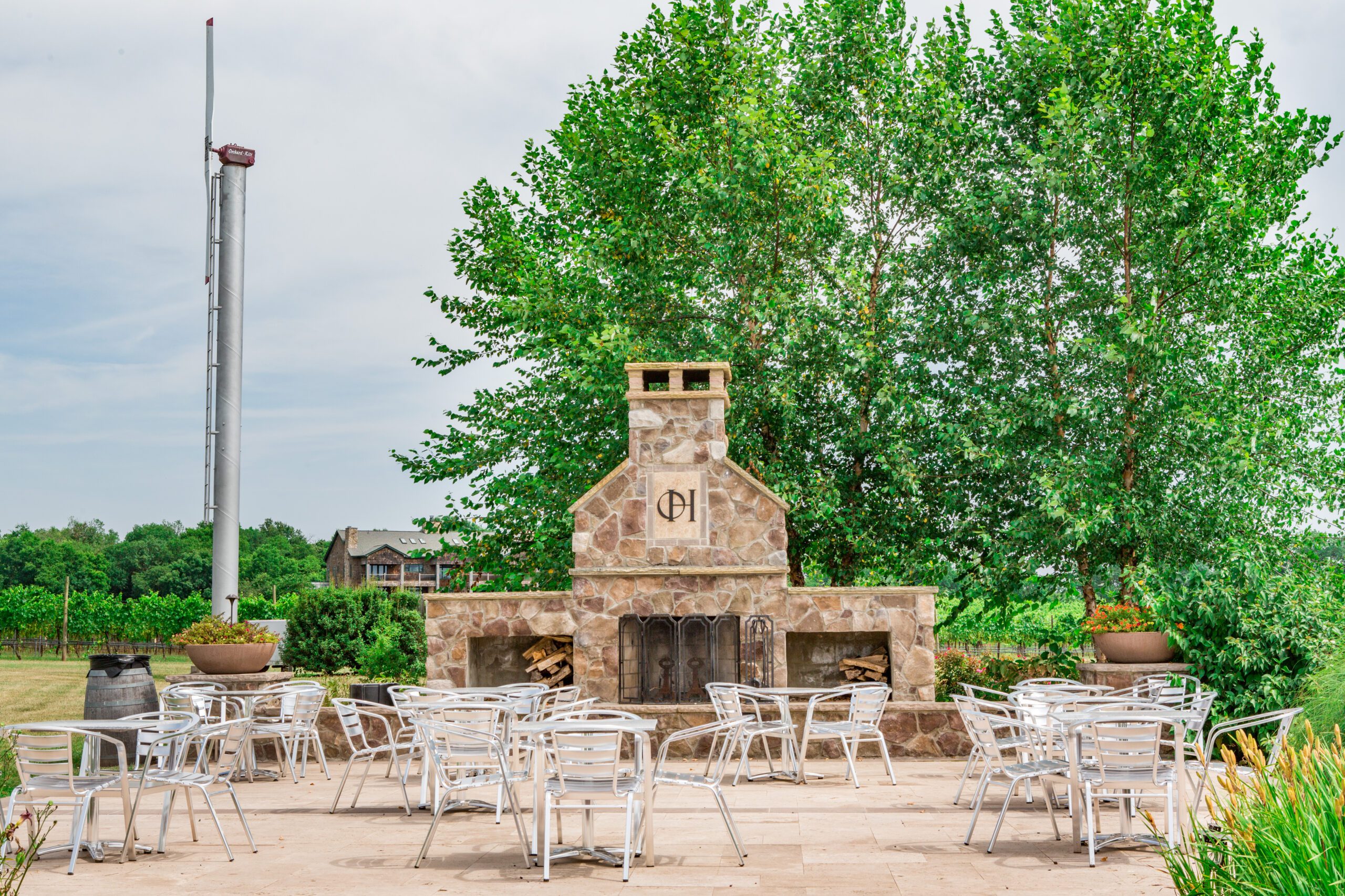An outdoor patio featuring a stone fireplace adorned with a circular emblem, surrounded by numerous white metal chairs and tables. Lush green trees stand in the background, with a tall flagpole visible on the left side. A few distant buildings can be seen.