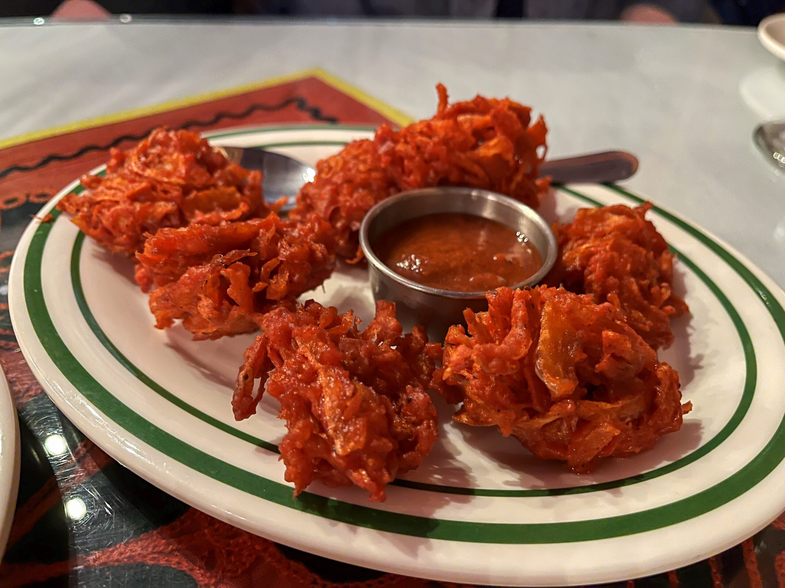A white plate with green stripes holds several pieces of crispy, deep-fried onion bhajis next to a small metal bowl containing a red dipping sauce. A spoon rests on the edge of the plate. The background shows a patterned tabletop.